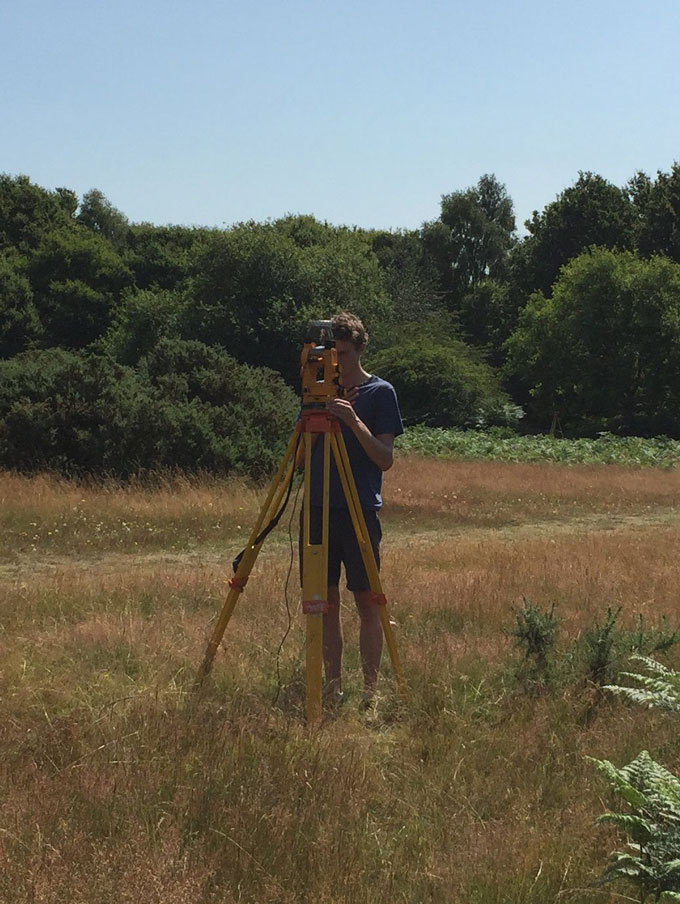 A man standing in a field of of yellow and green grass looking through a piece of equipment called a theodolite. The theodolite is standing on a yellow metal tripod. There are lush green trees and blue sky in the background.